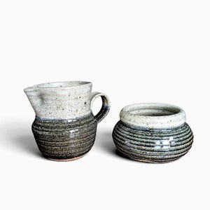 Peut inclure: Un pot à lait et un sucrier en céramique assortis présentant un design bicolore. Les parties supérieures sont blanc cassé moucheté, tandis que les corps inférieurs sont gris anthracite foncé texturé avec des détails nervurés horizontaux.