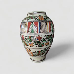 May include: A cream-colored ceramic vase with a round body and a narrow neck. The vase is adorned with colorful floral and geometric patterns in red, green, orange, and blue. The design includes stylized flowers and leaves, creating a folk art aesthetic.