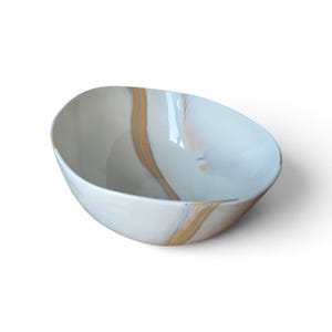 May include: A smooth, asymmetrical ceramic bowl featuring a glossy white finish with prominent, flowing vertical streaks of warm orange and soft grey marble-like patterns that wrap around the curved interior and exterior surfaces of the vessel.