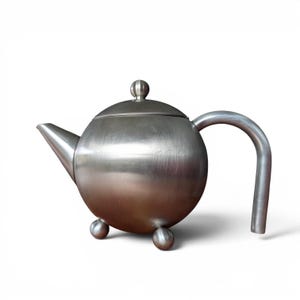May include: A brushed metal teapot with a spherical body, a curved handle, and a spout. The teapot has a small, round lid with a knob and rests on three small ball feet. The overall design is modern and minimalist.