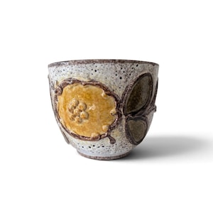 Peut inclure: Une tasse en céramique avec un émail blanc cassé moucheté présentant un motif de fleur jaune en relief sur le devant. Le bord et la base sont finis avec un contour brun foncé, et la surface extérieure affiche un aspect terreux et tacheté.