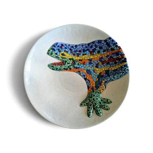 May include: A round, off-white ceramic plate featuring a raised, colourful mosaic lizard design. The lizard is composed of small, multi-coloured tiles in shades of blue, yellow, orange, green, and red, covering the head and one front foot.