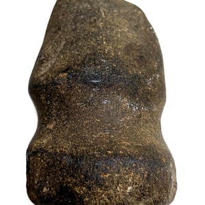Pode incluir: Um artefato de pedra marrom escuro e desgastado, com um topo arredondado e uma base alargada. A pedra tem uma superfície áspera e texturizada com áreas mais escuras, sugerindo idade e uso. A forma é aproximadamente retangular.