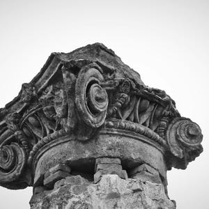 Puede incluir: Imagen en blanco y negro de un detalle arquitectónico ornamentado y desgastado. Se ve la parte superior de una columna, posiblemente de una estructura clásica. Presenta intrincadas tallas, incluyendo volutas en forma de espiral y motivos florales.