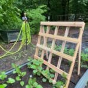 Könnte beinhalten: Ein Holzspalier lehnt an einem erhöhten Beet mit dunkler Erde und grünen Pflanzen. Ein gelber Gartenschlauch und ein Wassersprenger sind im Hintergrund zu sehen, mit grünen Bäumen in der Ferne. Das Spalier ist aus hellem Holz gefertigt.