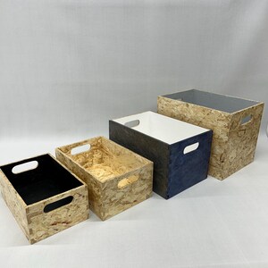 May include: Four wooden storage boxes of varying sizes. The boxes have a natural wood grain finish, with cut-out handles. The interiors are black, white, and navy blue. The boxes are arranged in a row, with the largest box in the back.