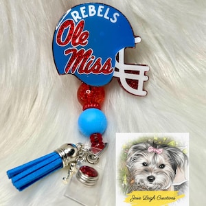 May include: A blue and red badge reel featuring a football helmet design with the words "Rebels" and "Ole Miss". The reel has a blue tassel, red and blue beads, and a small dog illustration with the text "Josie Leigh Creations".