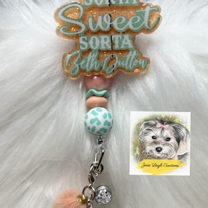 May include: A badge reel with the text "Sorta Sweet Sorta Beth Dutton" in a glittery design. It features a pink and teal bead design, a fluffy peach pom-pom, and a small charm. A small image of a dog is also included.