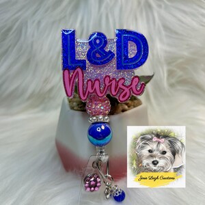 May include: A blue and pink L&D Nurse badge reel with a beaded design. The badge reel has the text "L&D Nurse" in blue and pink glitter. A small image of a dog is on the right side of the badge reel.