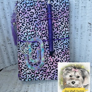 May include: A notebook cover with a purple and blue leopard print pattern, featuring a purple glitter pen, a large floral paperclip with a heart charm, and a purple tassel. A square card shows a dog illustration with the text Josie Leigh Creations.