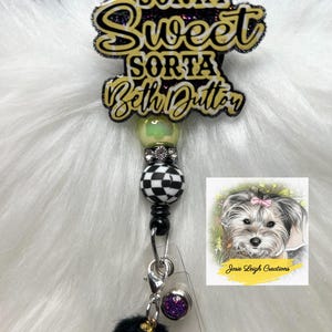 May include: A badge reel with the text "Sorta Sweet Sorta Beth Dutton" in gold and black. The reel has a black and white checkered bead, a black pom-pom, and a small image of a dog with a pink bow.