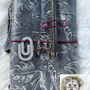 May include: A gray leather journal with a floral embossed design. The journal is secured with a burgundy elastic band and features a silver pen, a pink paperclip, and silver charms. A gray tassel hangs from the side.