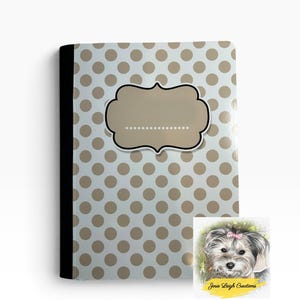 May include: A notebook with a white cover featuring a repeating pattern of tan polka dots and a black spine. Centered on the front is a tan label with a black border and a dotted line for writing. A small square logo shows a dog with a pink bow.