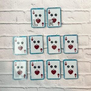 May include: Ten rectangular resin charms featuring a white ace of hearts playing card design with a cute cartoon face. Each card has a blue glitter border. Two of the top cards feature silver letter charms L and R respectively.
