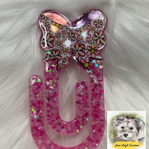 May include: A pink glittery resin bookmark with a bow-shaped top featuring a floral pattern in pink, red, and white. The bookmark has a U-shape and is filled with glitter. A small illustration of a dog is in the bottom right corner.
