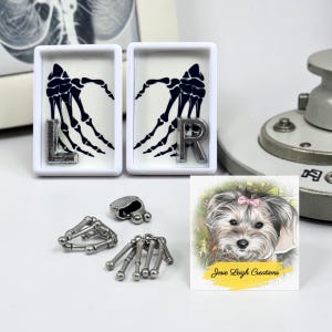 May include: Two white rectangular frames containing black skeletal hand illustrations with silver L and R letters, accompanied by small silver-toned metal bone-shaped charms and a square card featuring a dog illustration with the text Josie Leigh Creations.
