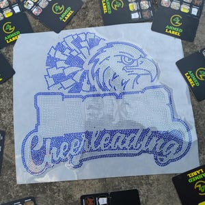 May include: A rhinestone design featuring an eagle and the words "EAGLES Cheerleading" in a blue and white color scheme. Several black cards with the "AHMED LABEL" logo are scattered around the design.