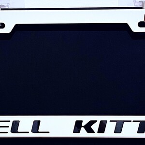 May include: White license plate frame with the words "HELL KITTY" in bold, black letters. The frame has a rectangular shape with rounded corners and a black background. Two small hooks are at the top.