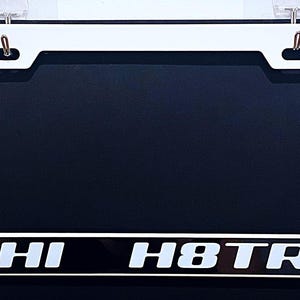 May include: White license plate frame with the text "HI H8TR" in white lettering on a black background. The frame has a rectangular shape with rounded corners and mounting holes at the top.