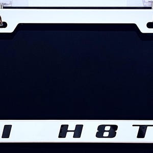 May include: White license plate frame with the text "HI H8TR" in black block letters. The frame has a rectangular shape with rounded corners and is designed to fit a standard U.S. license plate. The background is a dark blue.