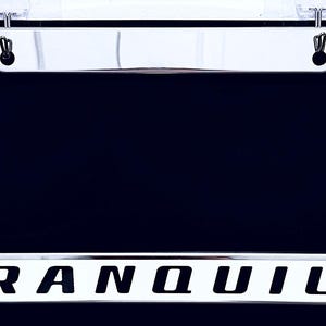 May include: A rectangular chrome license plate frame with a polished metallic finish. The bottom horizontal bar features a white rectangular insert with the word TRANQUILO printed in bold, black, sans-serif capital letters. Two mounting holes.