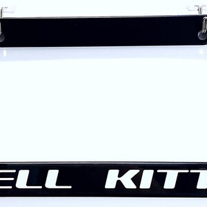 May include: Black license plate frame with the words "HELL KITTY" in white block letters. The frame has a rectangular shape with rounded corners and is designed to hold a standard license plate. Includes mounting hardware.