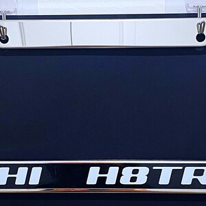 May include: A shiny chrome license plate frame with the words "HI H8TR" in white lettering on a black background. The frame has a rectangular shape with rounded corners and mounting holes at the top.