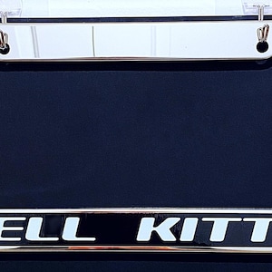 May include: A chrome license plate frame with the words "HELL KITTY" in white block letters. The frame has a shiny, reflective surface and is mounted on a dark blue background.
