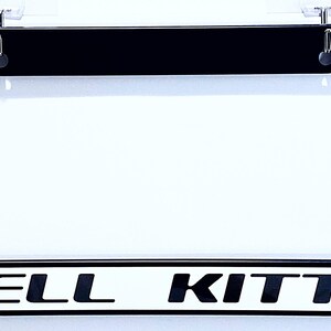 May include: Black license plate frame with the words "HELL KITTY" in black block letters on a white background. The frame is rectangular and has a clear plastic cover.