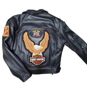 May include: A black leather motorcycle jacket featuring a large embroidered patch on the back with an eagle and the Harley-Davidson Motor Cycles logo. Above the eagle is a POW-MIA patch with the text ARE NOT FORGOTTEN. A Marine Corps patch is on the sleeve.