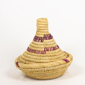 May include: A woven basket with a conical lid. The basket is made of natural-coloured straw with bands of purple detailing. The base is round, and the lid tapers upwards. The basket is likely used for storage or decoration.