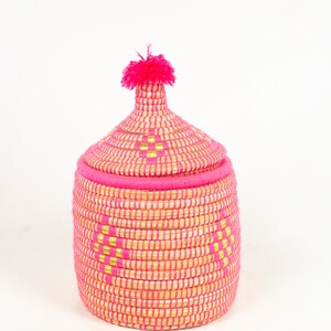 May include: A round, lidded basket in a vibrant pink and yellow woven pattern. The basket features a decorative pink tassel on top of the lid. The design includes a repeating geometric pattern in gold. The basket is suitable for storage or decoration.
