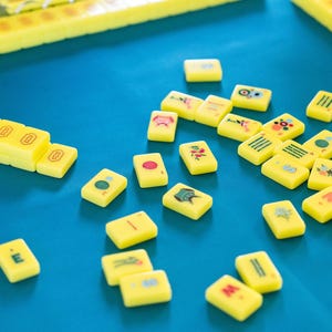 May include: A collection of yellow Mahjong tiles with various symbols and characters scattered on a blue surface. Several yellow tile racks are also visible. The game pieces are brightly colored and arranged for play.