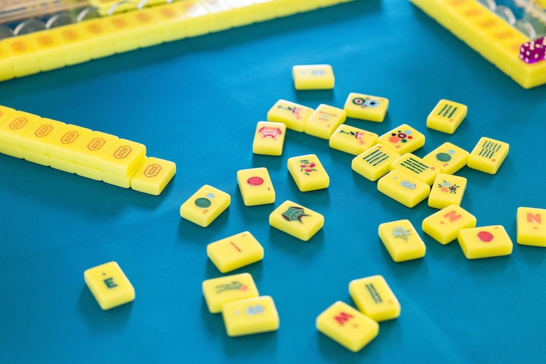 May include: A collection of yellow Mahjong tiles with various symbols and characters scattered on a blue surface. Several yellow tile racks are also visible. The game pieces are brightly colored and arranged for play.
