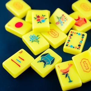 May include: A collection of yellow Mahjong tiles with various colorful illustrations and symbols. Some tiles feature floral designs, while others display numbers, lines, and other unique graphics. The tiles are scattered on a dark blue surface.