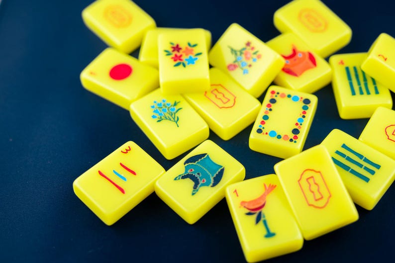 May include: A collection of yellow Mahjong tiles with various colorful illustrations and symbols. Some tiles feature floral designs, while others display numbers, lines, and other unique graphics. The tiles are scattered on a dark blue surface.