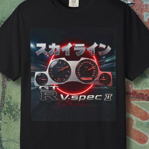 Könnte beinhalten: Schwarzes T-Shirt mit einem Auto-Armaturenbrett-Grafik, japanischem Text "Skyline" und "GT-R V-spec II". Das Armaturenbrett ist silber und rot, vor dunklem Hintergrund.
