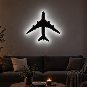 Peut inclure: Une silhouette noire d'avion commercial fixée au mur, dotée d'un rétroéclairage LED blanc brillant qui crée un effet de halo lumineux autour des bords de la forme de l'appareil, soulignant ses ailes, son fuselage et sa section de queue.
