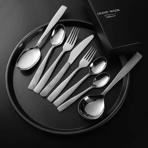Puede incluir: Un juego de cubiertos de acero inoxidable, que incluye cucharas, tenedores y un cuchillo, dispuestos sobre una bandeja negra. Los mangos tienen un patrón de diamantes texturizado. Se ve una caja negra con el texto "GRAND WAZIR Tableware".