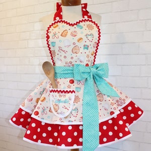 Puede incluir: Un delantal blanco con la parte superior en forma de corazón y la parte inferior con lunares rojos y blancos. El delantal tiene un lazo turquesa y un bolsillo con el nombre "Isabelle" bordado. El delantal está decorado con un tema navideño, incluyendo bastones de caramelo, copos de nieve y galletas de jengibre.
