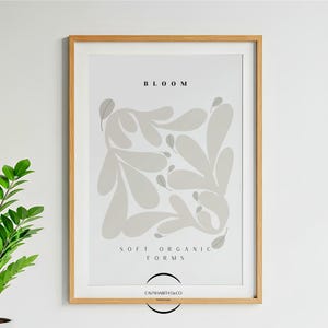 Puede incluir: Una impresión enmarcada con un diseño minimalista. La obra de arte presenta formas orgánicas suaves en tonos grises sobre un fondo blanco. La palabra "BLOOM" está en la parte superior, con "SOFT ORGANIC FORMS" debajo. El marco es de color madera clara.