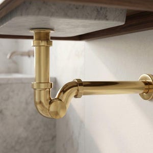 Op de afbeelding: Een goudkleurige metalen sifon geïnstalleerd onder een marmeren wastafel. De leidingconstructie bestaat uit een verticale afvoerbuis verbonden met een gebogen sifongedeelte en een horizontale buis die naar de muur loopt met een ronde flens.