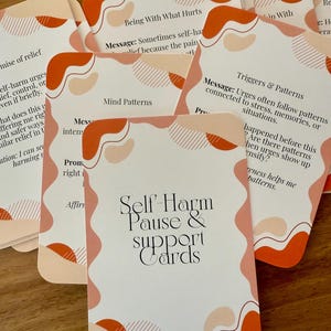 Könnte beinhalten: Ein Kartenspiel "Self-Harm Pause & Support Cards" mit weißem Hintergrund und abstrakten orangefarbenen und rosa Formen. Die Karten enthalten Texte wie "Being With What Hurts" und "Triggers & Patterns". Die Karten liegen auf einer Holzoberfläche.