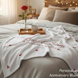 Puede incluir: Una manta blanca con detalles de corazones rojos y los nombres "Lucy & Sam" y la frase "Forever & Always" impresos. La manta está sobre una cama con una bandeja con una taza y una rosa en un jarrón.