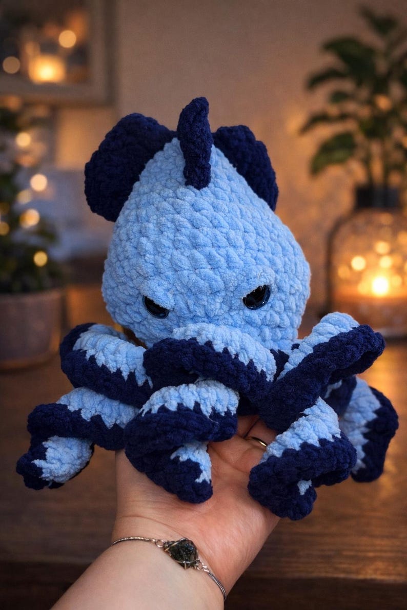 May include: A handmade, light blue and navy blue crocheted octopus plush toy. The octopus has dark eyes and a textured, soft appearance. The tentacles are a darker blue with light blue accents. The toy is designed to be a cuddly, decorative item.