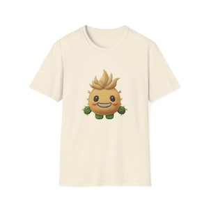 Puede incluir: Camiseta de color crema con un personaje de cactus de dibujos animados con una cara sonriente, pelo puntiagudo y brazos y pies de cactus verdes. El gráfico está centrado en la parte delantera de la camiseta.
