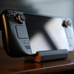 Peut inclure: Une console de jeu portable noire au design épuré, posée sur un support en bois. La console comprend un joystick, des boutons et un écran, mettant en valeur une esthétique moderne. L'appareil est conçu pour le jeu.