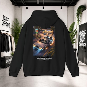May include: A black hooded sweatshirt featuring a rectangular graphic on the back. The print depicts an alien driving a silver sports car at a gas station at night. Below the image, the text reads A.M.F. STUDIO UNCHARTED DESIGNS 1984.