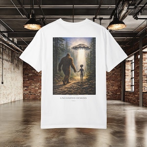Puede incluir: Camiseta blanca con un diseño gráfico colorido de un Bigfoot tomados de la mano con un alienígena bajo un OVNI. La escena se desarrolla en un bosque con un rayo de luz. El texto "UNCHARTED DESIGNS 1984" está impreso debajo.