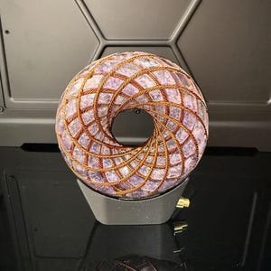 May include: A donut-shaped sculpture with a central opening. The sculpture is made of small, irregular purple stones held together by a woven copper wire. It sits on a black base, reflecting on a glossy black surface.